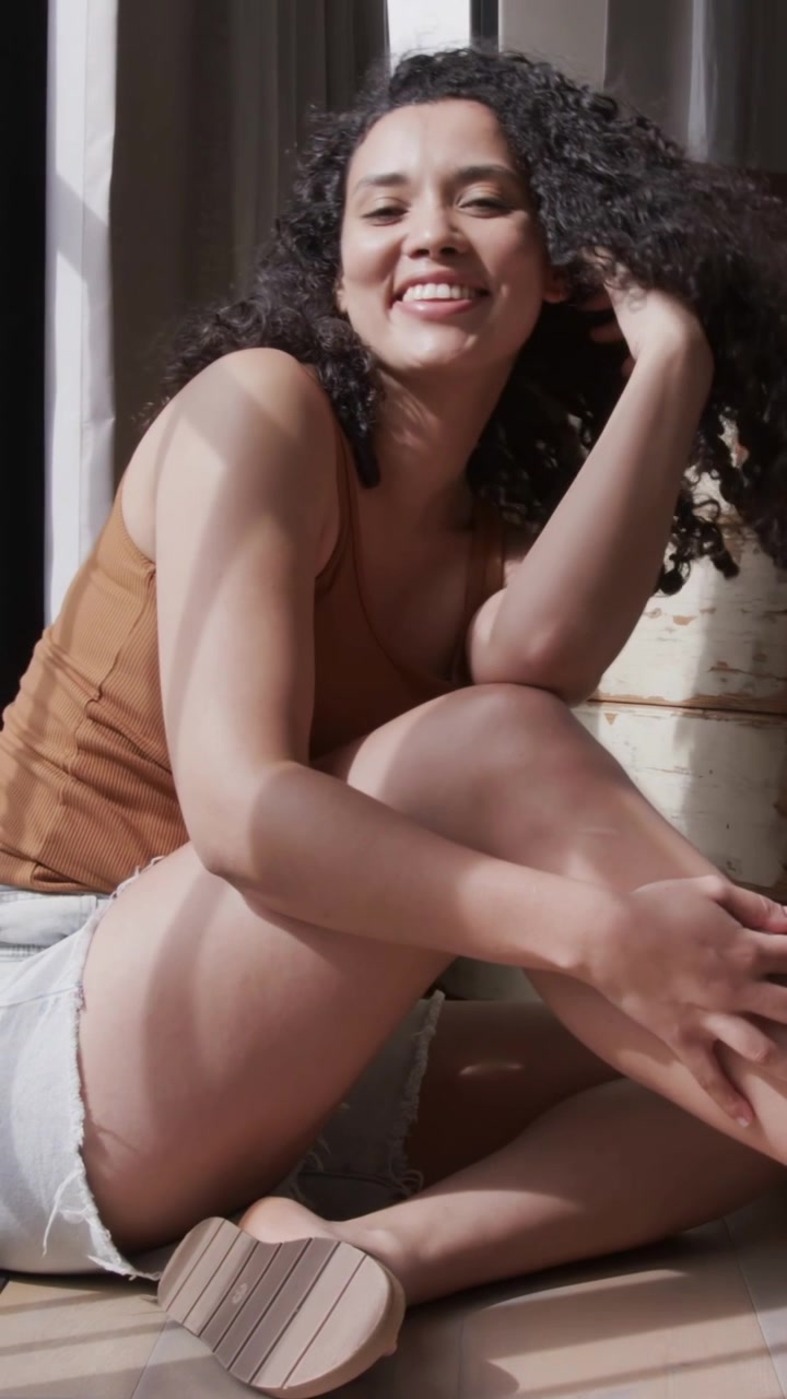 Video verticale: La luce delle persiane illumina una signora sorridente seduta vicino alla finestra, con canotta marrone, pantaloncini di jeans e un baule.