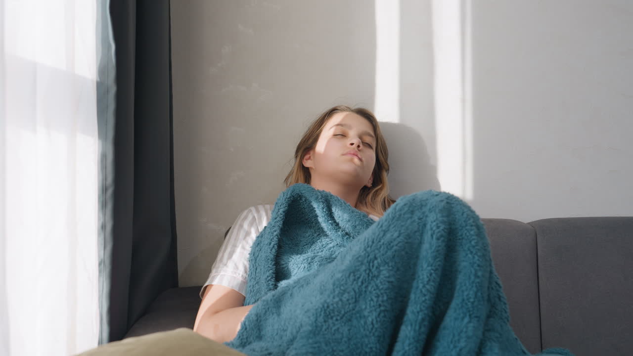 Adolescente si gode un tranquillo riposo diurno in casa, ragazza con coperta blu che fa un pisolino rilassato al chiuso, adolescente che dorme serenamente sul divano con la luce del sole che proietta strisce sul suo viso