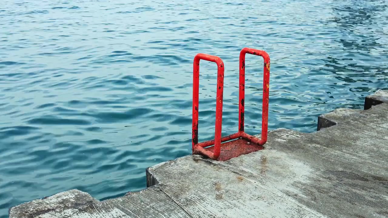 Red railing near water edge at a dock on a clear day showing calm waves and concrete surface