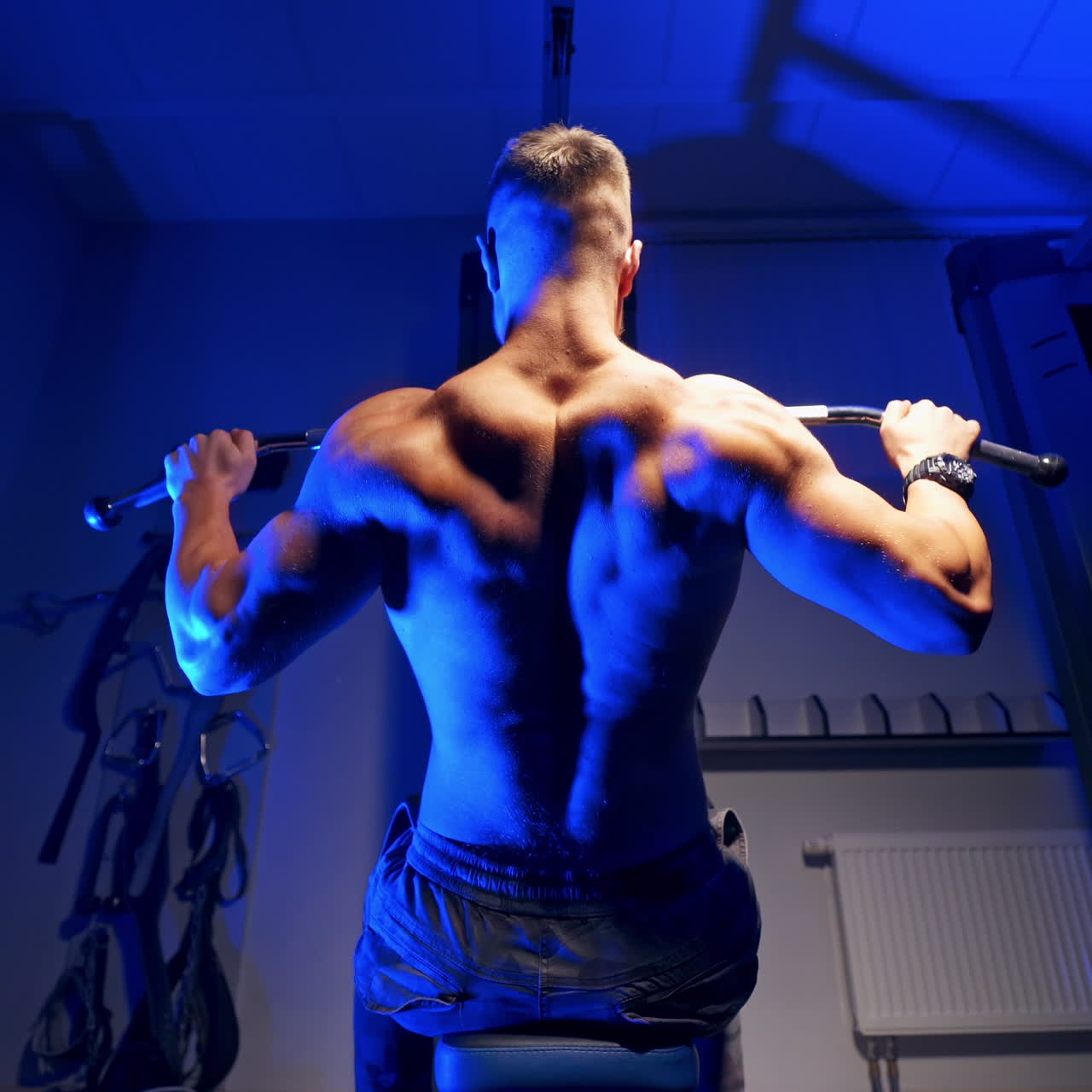 Backside view of an athlete exercising. Sportsman sitting and doing his workout on a simulator in the sports center. Blue light.