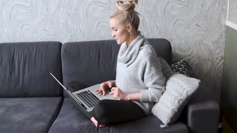 Cheerful-young-blond-woman-sitting-on-couch-in-living-room-and-using-laptop