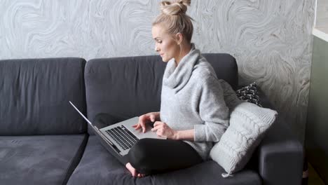 cheerful young blond woman sitting on couch in living room and using laptop