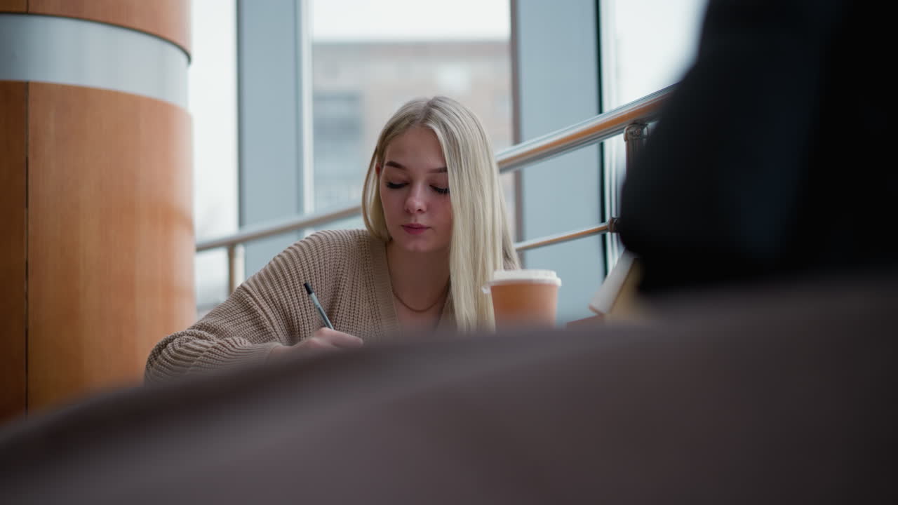 En ung kvinde skriver eftertænksomt i et indkøbscenter, med en kaffekop på et træbord, delvist udsyn til en uklar genstand i baggrunden, en afslappet og fredelig stemning.