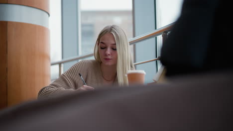 young woman thoughtfully writing in mall setting with coffee cup on wooden table, partial view of unclear object in background, casual and peaceful ambiance