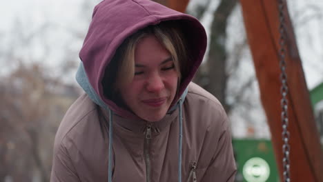 young girl in winter outfit bends slightly, showing discomfort, with a blurred snowy background and bare trees, hooded jacket covers head, facial expression conveys pain or distress in cold