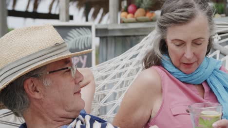 Happy-senior-caucasian-couple-drinking-and-talking-on-hammock-outside-beach-bar,-in-slow-motion