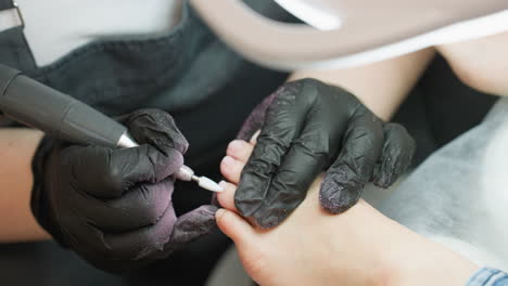 upward angle view showing nail technician wearing black gloves carefully filing client nail using electric tool under bright magnifying lamp with both hands steady in professional salon environment