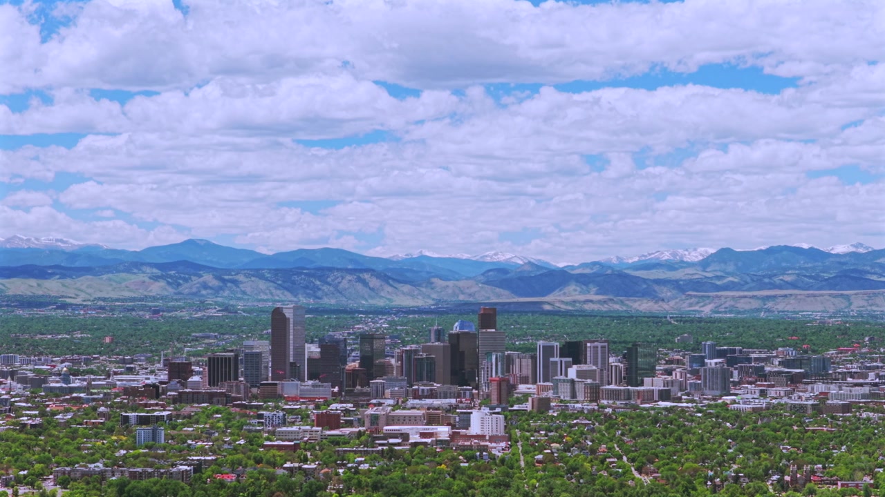 Drone luftfoto af Denver, Colorado, med byens skyline, skyskrabere og de sneklædte Rocky Mountains (Front Range) i baggrunden. Forårsmorgen med blå himmel og skyer over downtown Denver, med bevægelse mod venstre.