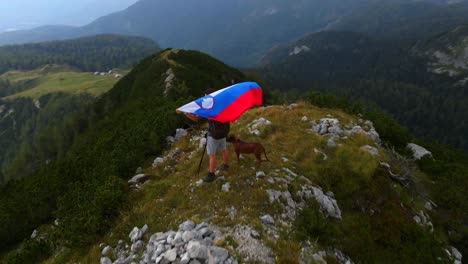 forward drone footage with a drone passing by a man holding a slovenia flag on top of a mountain in the alps filmed in 4k in nature and surrounding mountains