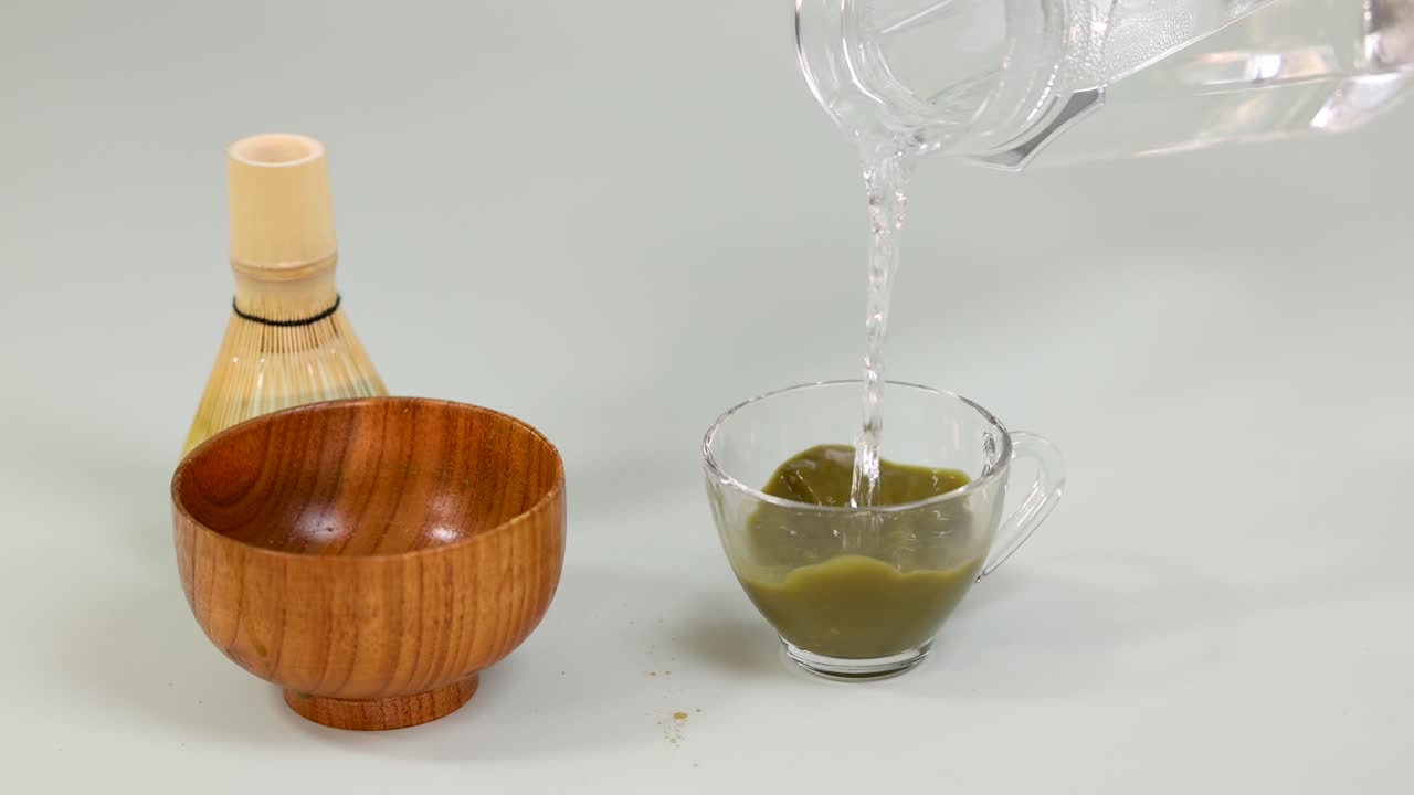 Hot water is poured into a glass cup with matcha powder, minimalistic neutral background