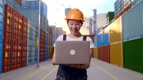 trabajadora asiática con gafas de protección y casco de seguridad sonriendo y señalando una computadora portátil en su mano mientras está de pie en el almacén del patio de contenedores