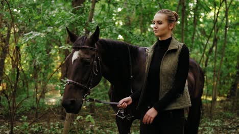 Attractive-young-female-jockey-is-walking-with-brown-horse-with-white-spot-on-forehead-in-park-during-sunny-day-holding-leather