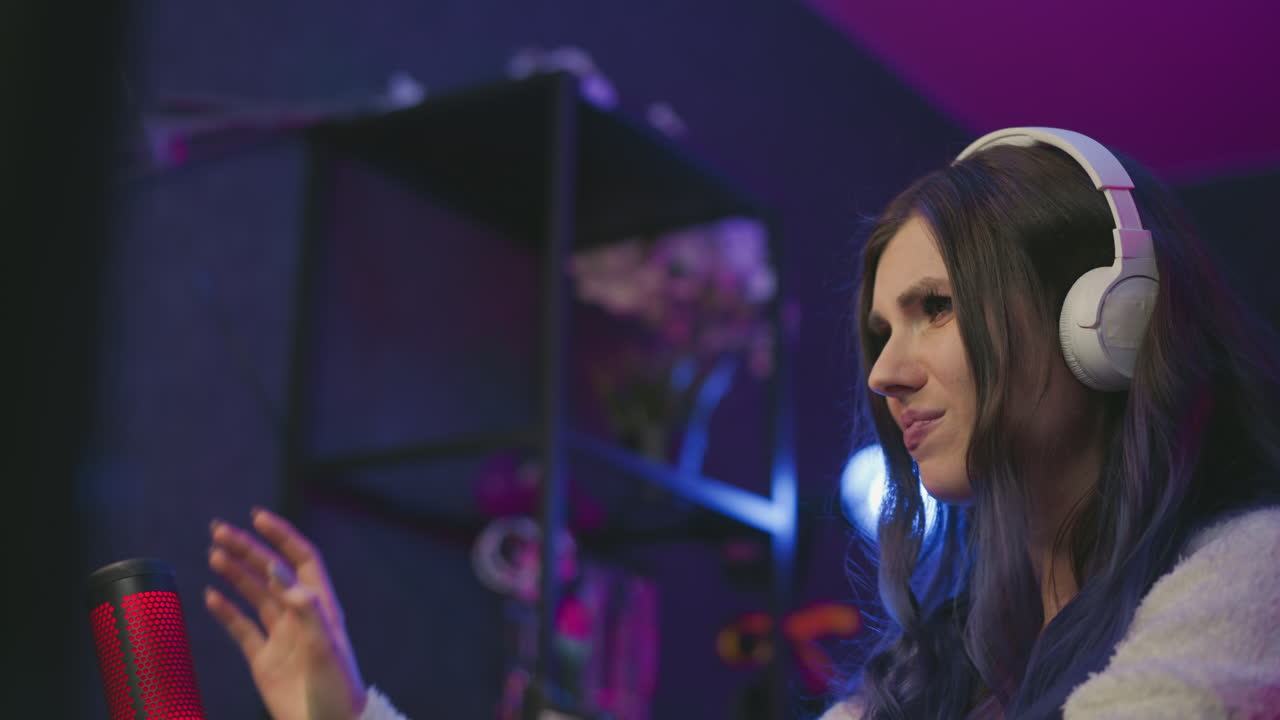 Curious female gamer wearing white headphones reacts with visible displeasure after losing game, seated in dimly lit neon studio with condenser microphone in front