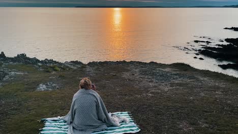 aerial of young hipster woman sitting on blanket in front of atlantic ocean in france, brittany taking photo during sunset golden hour