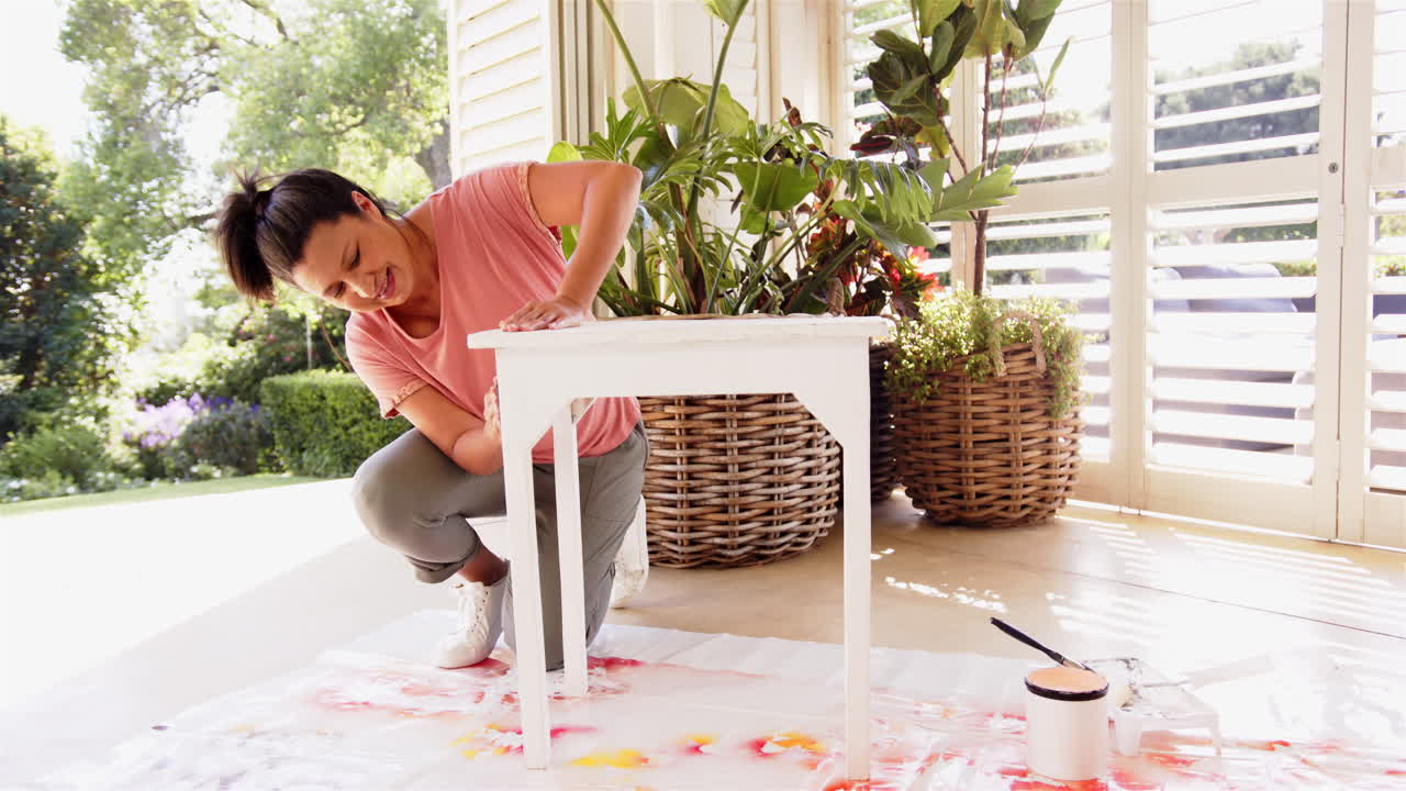 Painting white table, woman kneeling on floor with paintbrush and plants nearby