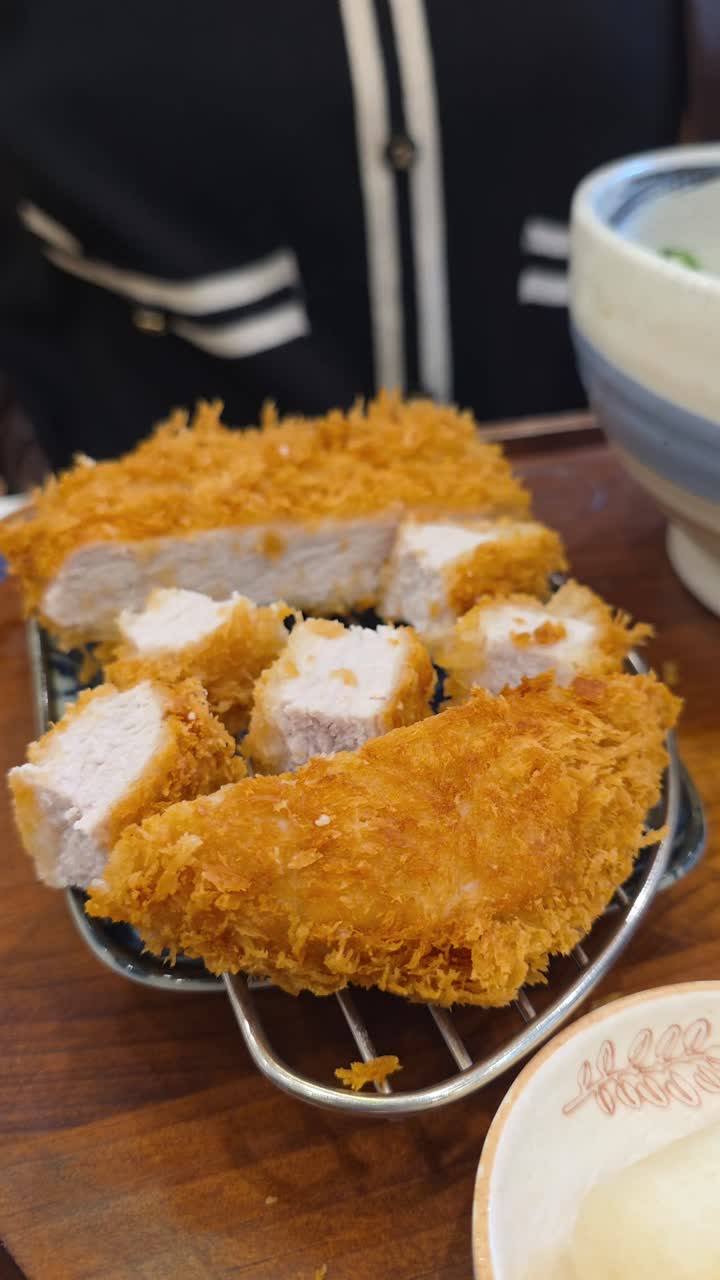 A close-up woman's hands using kitchen scissors and tongs to cut a crispy, deep-fried Japanese pork cutlet (tonkatsu) into bite-sized pieces before eating it in a restaurant