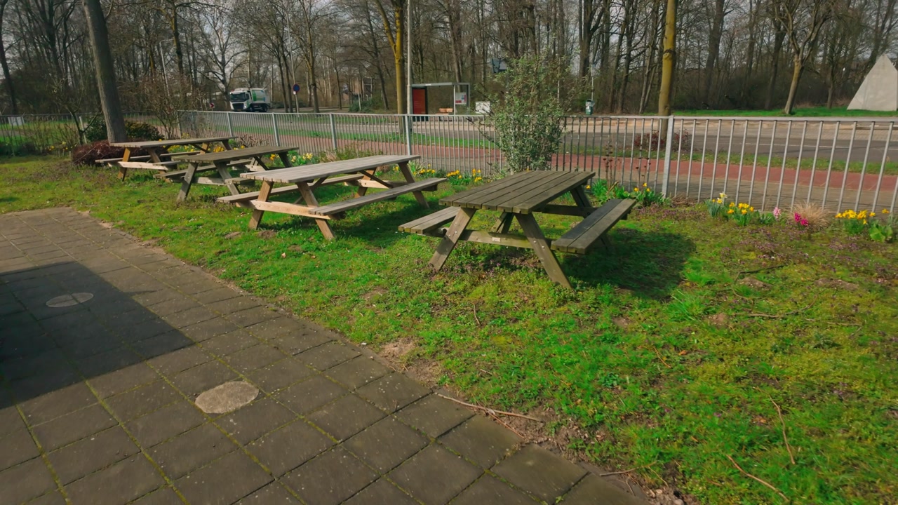 Tranquil outdoor picnic area featuring multiple rustic wooden tables and benches on a lush green lawn. Area is lined with early spring flowers, a paved path, and fence beside road. Bare trees visib...
