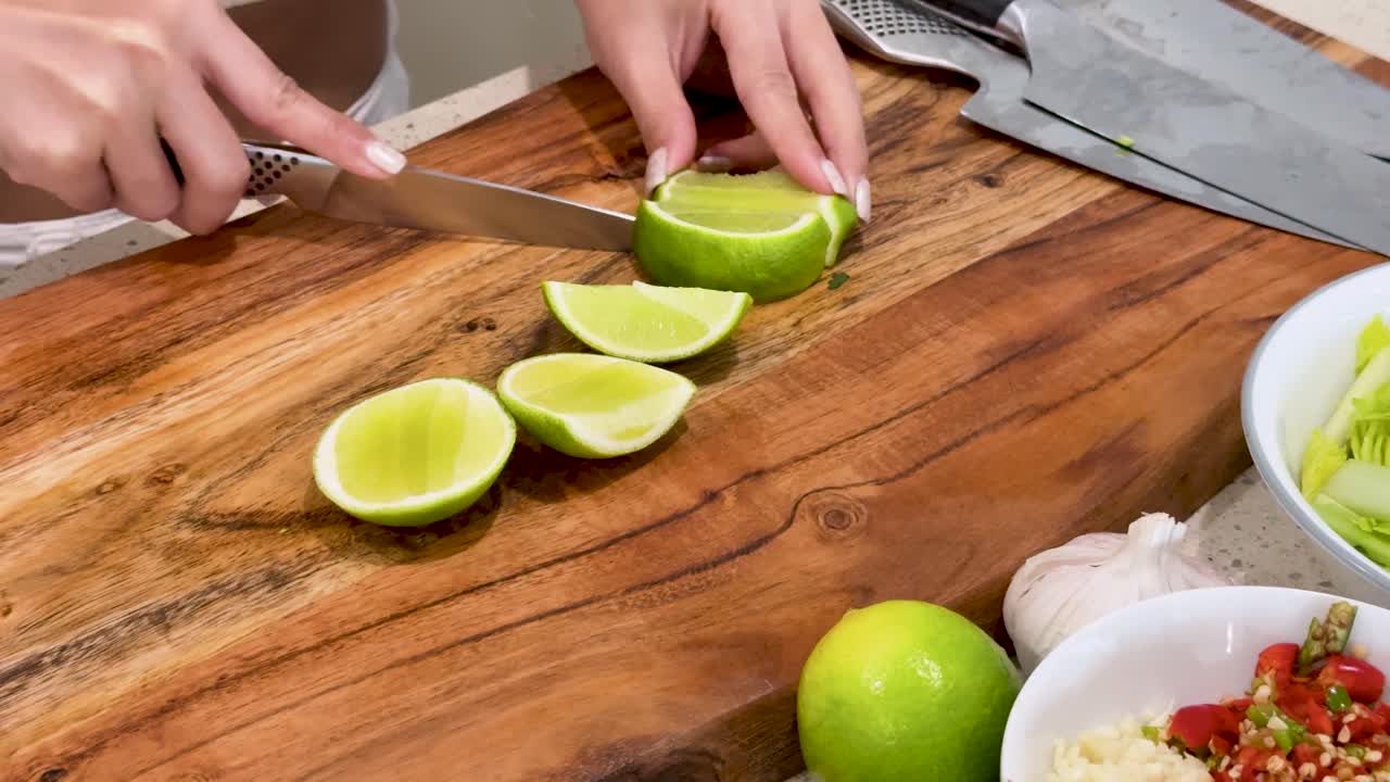 A woman uses a kitchen knife to cut a lime into wedges on a wooden cutting board in a brightly lit kitchen, surrounded by fresh ingredients