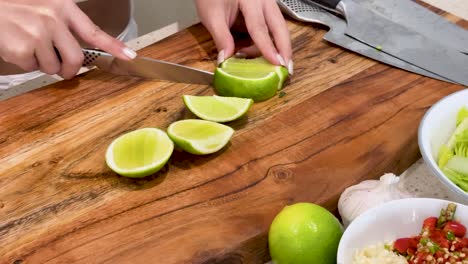 a woman uses a kitchen knife to cut a lime into wedges on a wooden cutting board in a brightly lit kitchen, surrounded by fresh ingredients