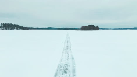 Volando-A-Lo-Largo-De-Una-Carretera-Sobre-El-Hielo-De-Un-Lago-Congelado-Para-Revelar-Un-Bosque-Nevado-En-El-Horizonte