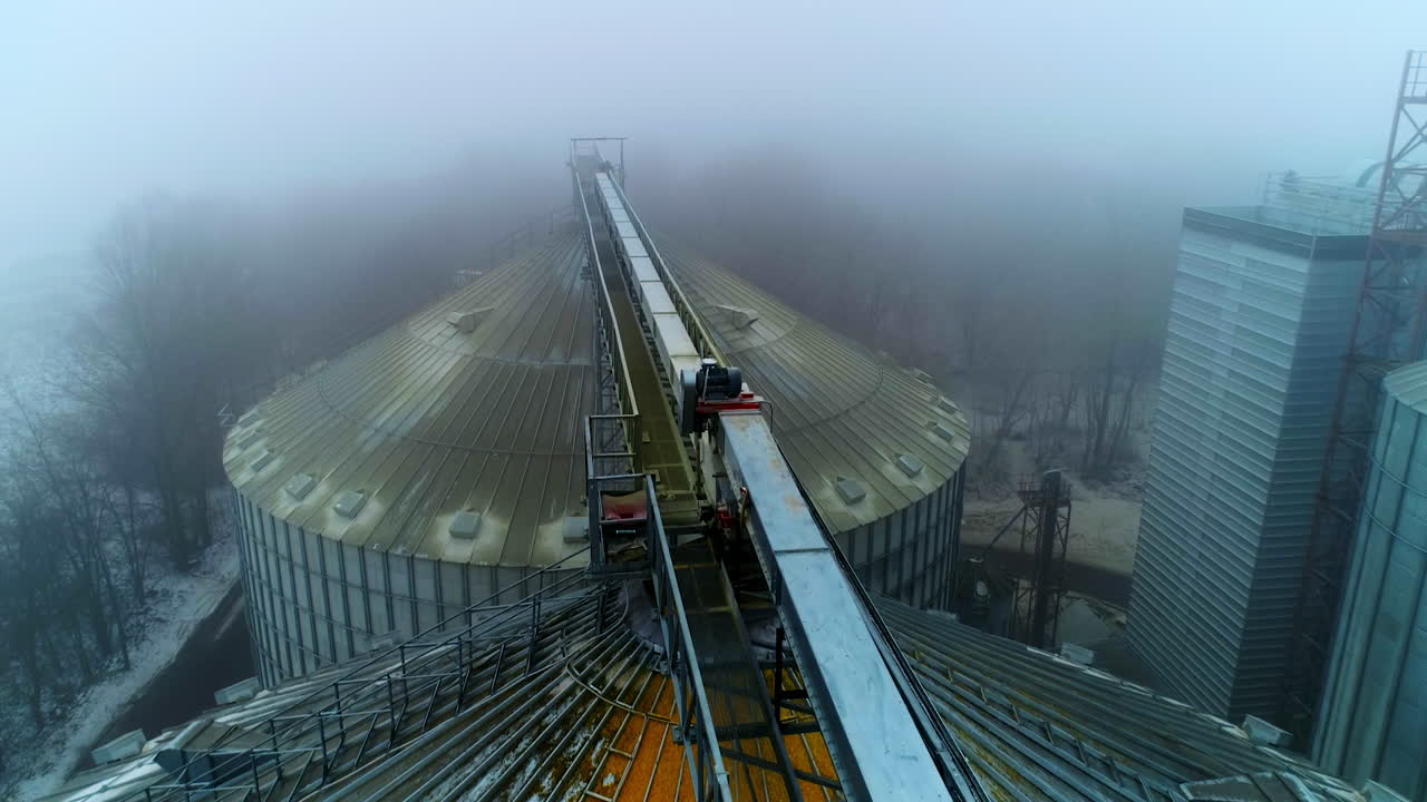Dusty tops of the bin at grain elevator complex. Drone footage above the metal tanks at modern agricultural plant. Foggy weather backdrop.