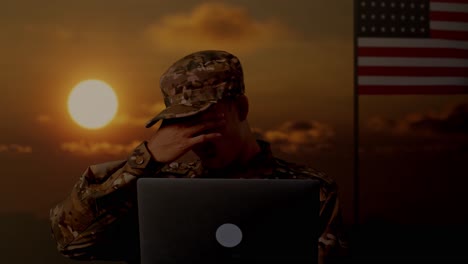 primer plano de un soldado asiático que usa una computadora portátil y tiene dolor de cabeza mientras está de pie con la bandera de los estados unidos, hora del atardecer