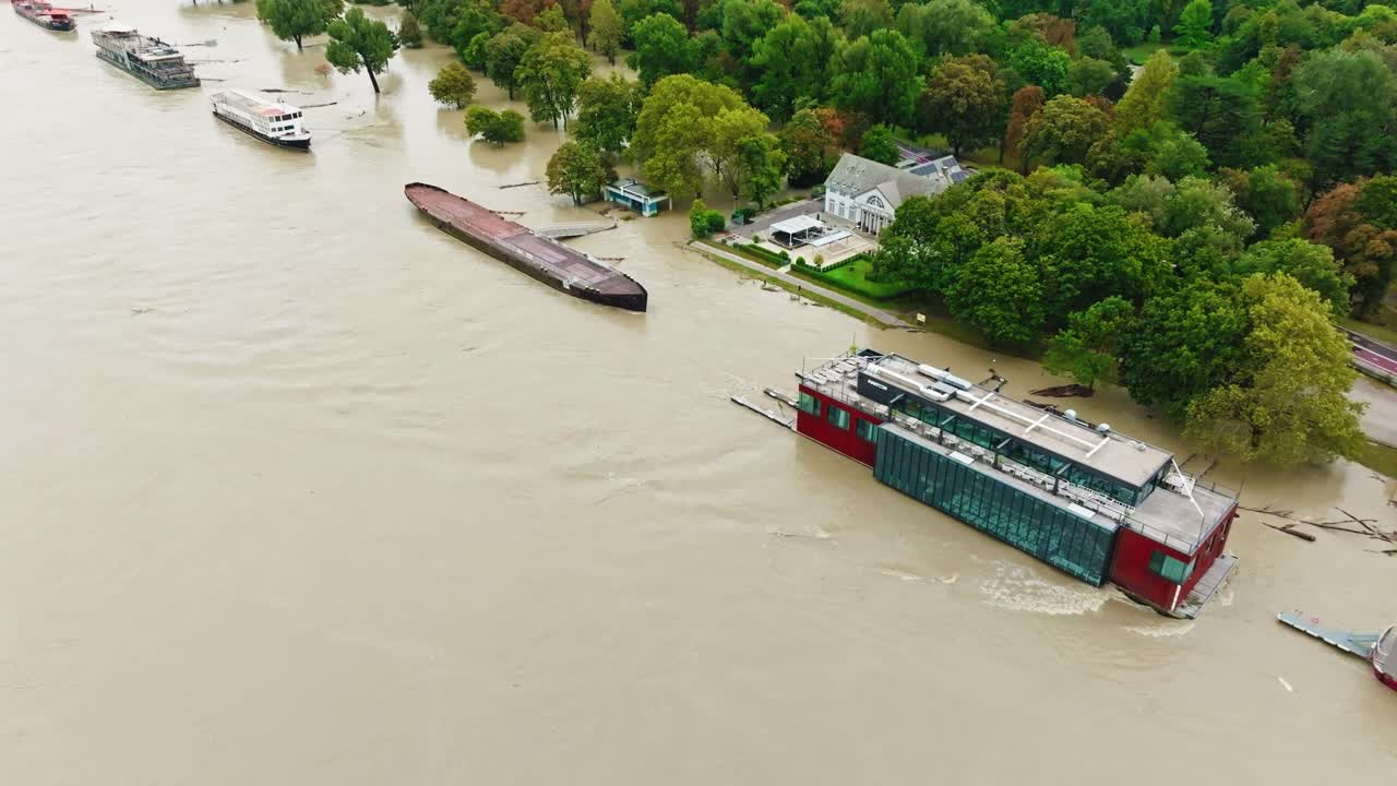 Aerial footage of the Danube River overflowing its banks in Bratislava, submerging the riverbank and surrounding trees. Boats and floating structures are caught in rising floodwaters.