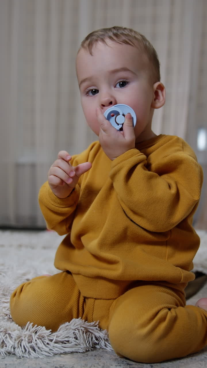 Pretty little baby sitting on the floor with pacifier in mouth. Kid looses the soother and picks it up and pulls to mouth. Vertical video