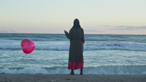 a woman holding a red balloon looks at the sea waves crashing