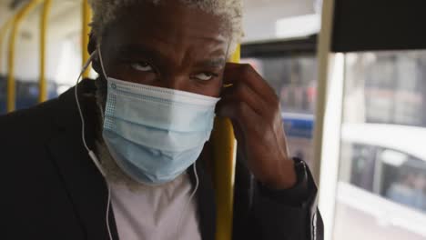 Afroamerikanischer-älterer-Mann-Mit-Gesichtsmaske-Und-Kopfhörern,-Während-Er-Im-Bus-Steht