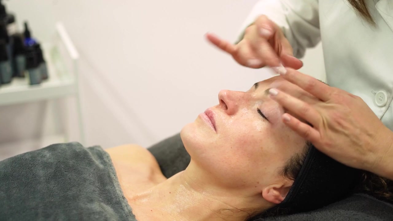 Cosmetologista massageando o rosto de uma jovem cliente com máscara cosmética em um salão de beleza.