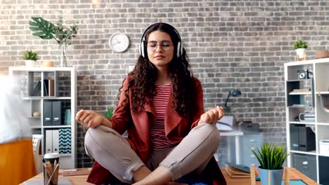 zoom fuera de la dama escuchando música relajándose en la oficina en la mesa en la postura del loto