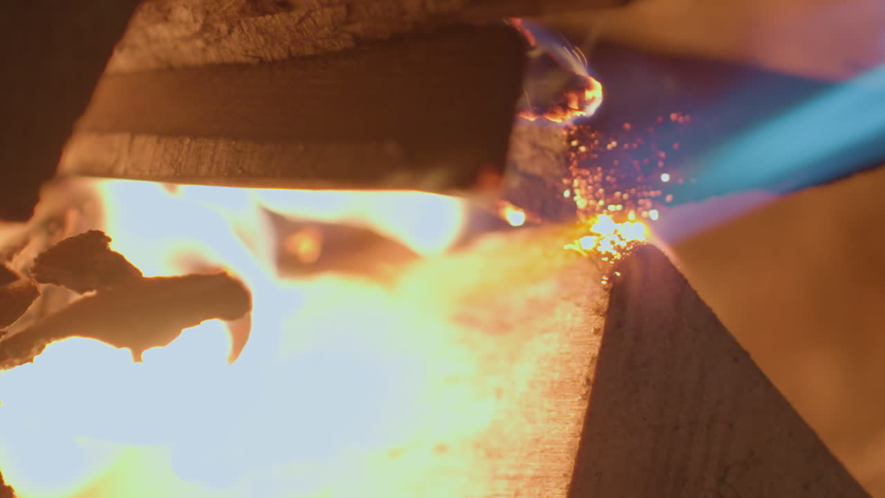 Extreme close up of person using blue gas torch to ignite stacked firewood inside dark fireplace, wood catches flame gradually, glowing and producing soft flickering light in warm indoor setting