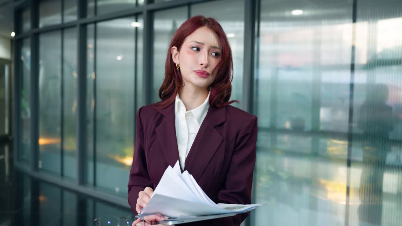 モダンなオフィス通路に立つ、心配そうで落胆した様子の若い女性幹部。大量の紙の報告書を、過重な仕事量に切羽詰まった表情で調べている。