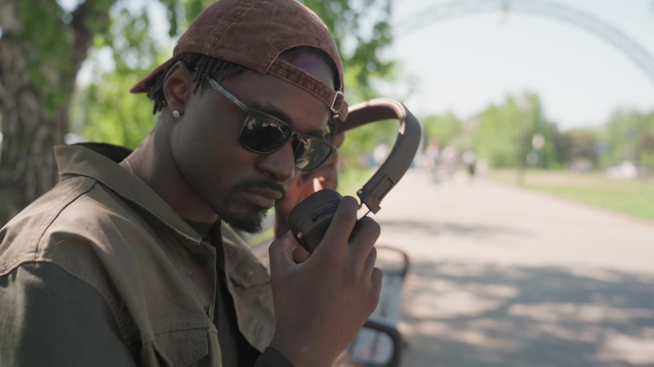 Close up of man wearing brown cap and headphones adjusting them while sitting on bench in park. Man is dressed in casual outdoor clothing with sunlight filtering through trees creating