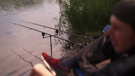 Fisherman-sitting-and-waiting-beside-fishing-rods-on-riverbank