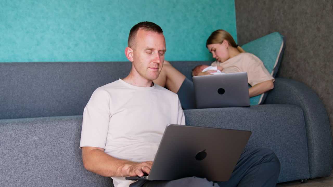 Busy mom of a newborn has to take care of a baby and do work. Man sitting at the sofa is fully focused on his work on laptop and asks his wife about issues on computer.