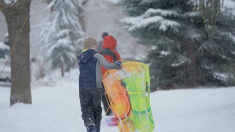 young white male walking on a snow covered sidewalk carrying a colorful sled