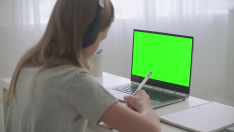 una adolescente está aprendiendo en línea sentada en la mesa frente a una computadora portátil con una tecla de croma en la pantalla escribiendo en un cuaderno en casa