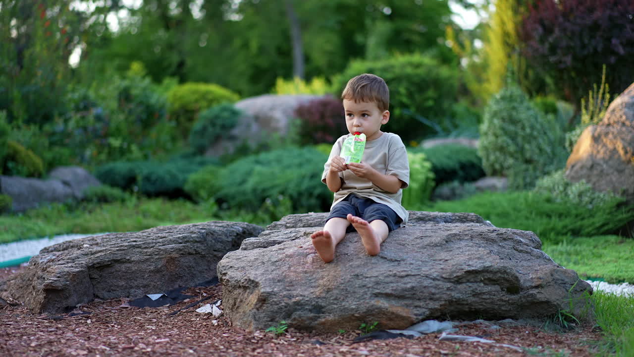 Sweet barefoot boy sitting outdoors on the rock holding a doy pack. Happy child drinking or eating from pack.