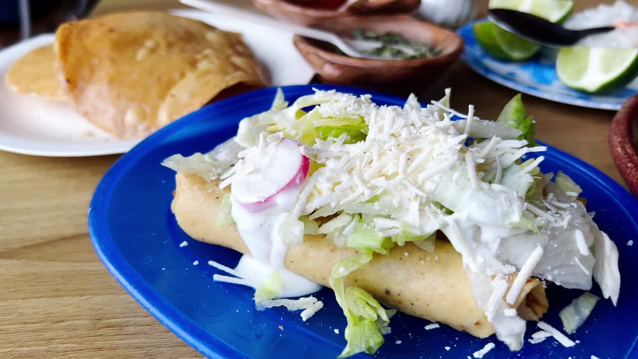 Two delicious flautas on a blue plate, covered with shredded cheese, cream, lettuce and radish slices, ready to be enjoyed with some tortillas and a side of green sauce, slow motion reveal