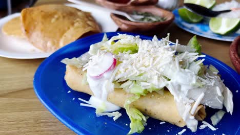 two delicious flautas on a blue plate, covered with shredded cheese, cream, lettuce and radish slices, ready to be enjoyed with some tortillas and a side of green sauce, slow motion reveal