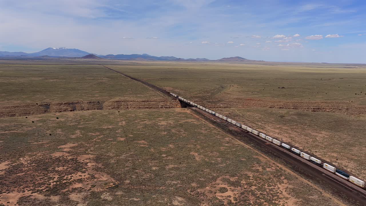 A high-angle drone aerial view captures a long freight cargo train moving through the expansive and arid southwest desert landscape under a clear sky