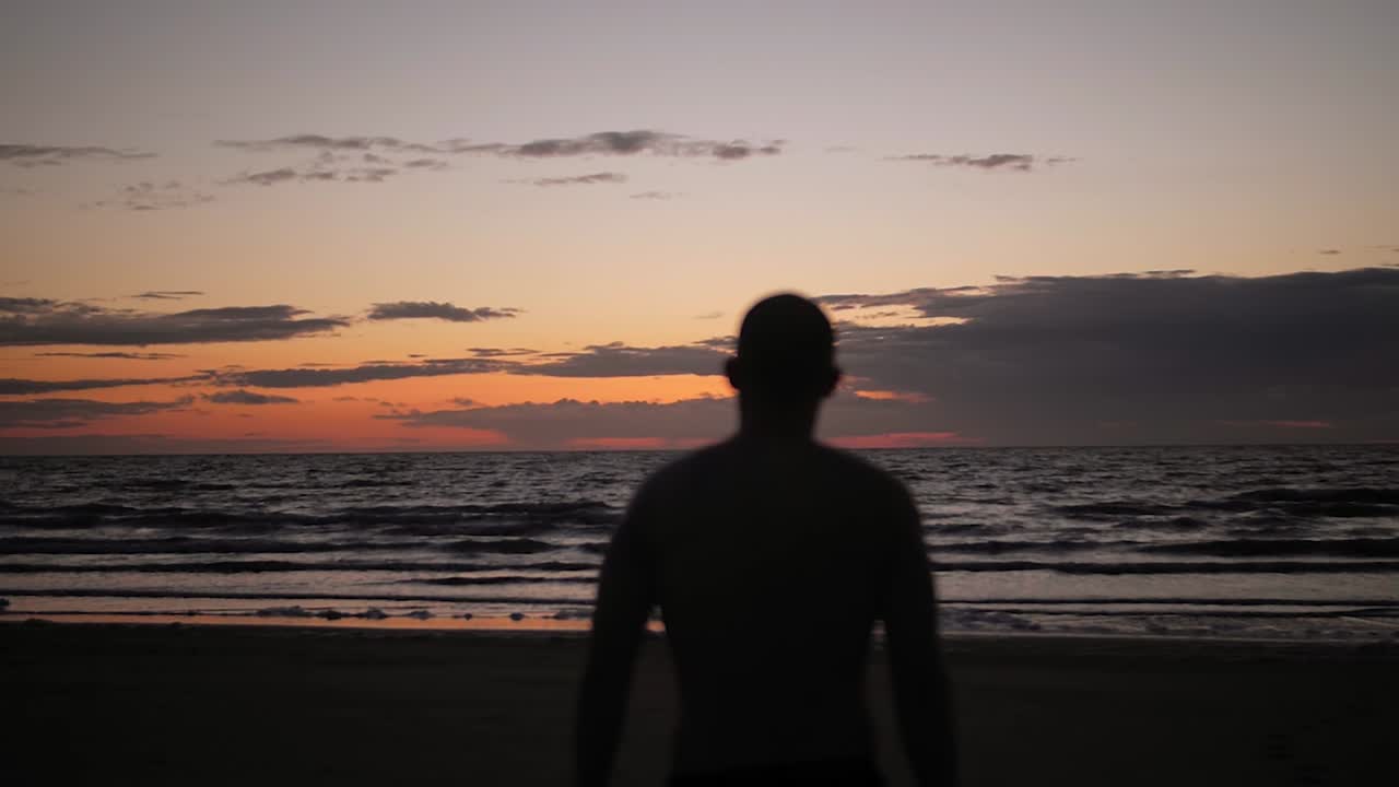 Sagoma di uomo che cammina verso il mare calmo all'alba al rallentatore, simboleggiando riflessione, resilienza e salute mentale.