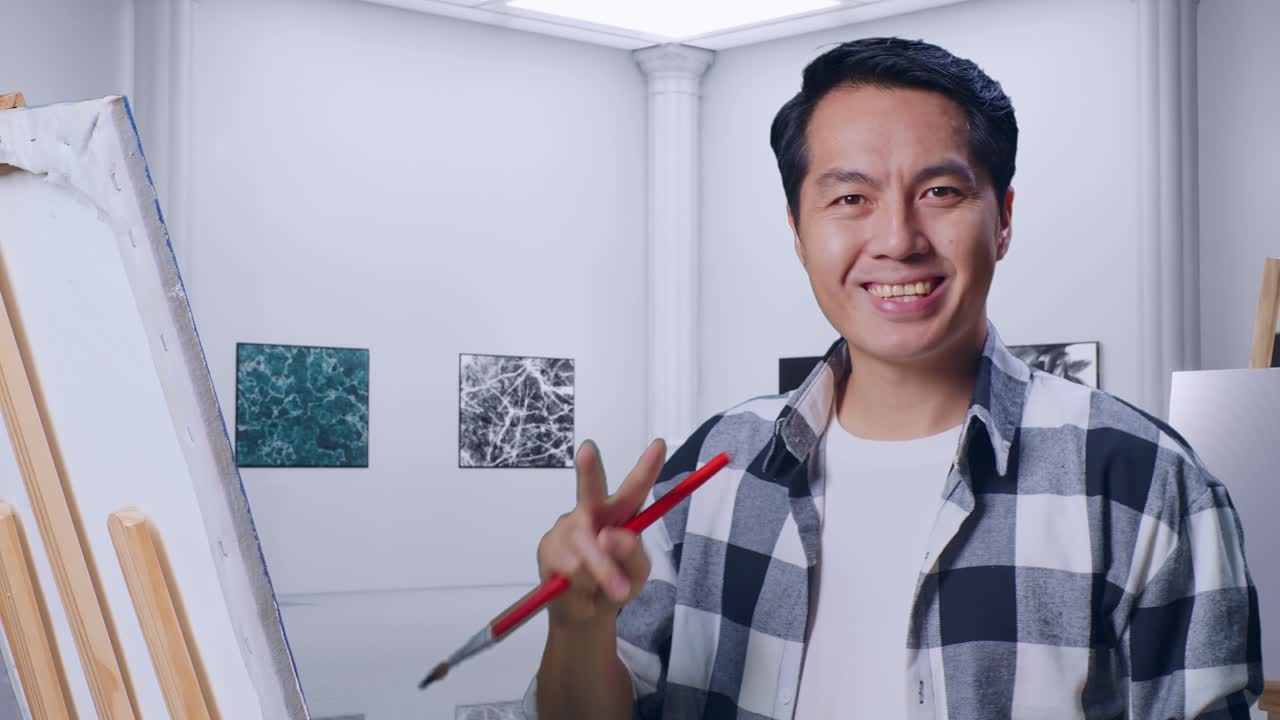 アジアの男性アーティストが笑顔でカメラに向かって平和のジェスチャーを示し画廊で油絵とブラシでキャンバスに絵を描いています