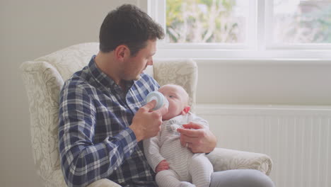 Father-feeding-baby-son-with-bottle-sitting-in-chair-at-home-together---shot-in-low-motion