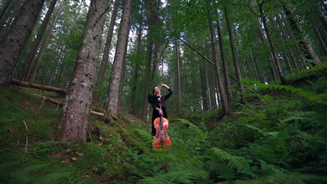 pretty woman with cello in forest. beautiful young female musician standing inside the green woods and holds musical instrument.