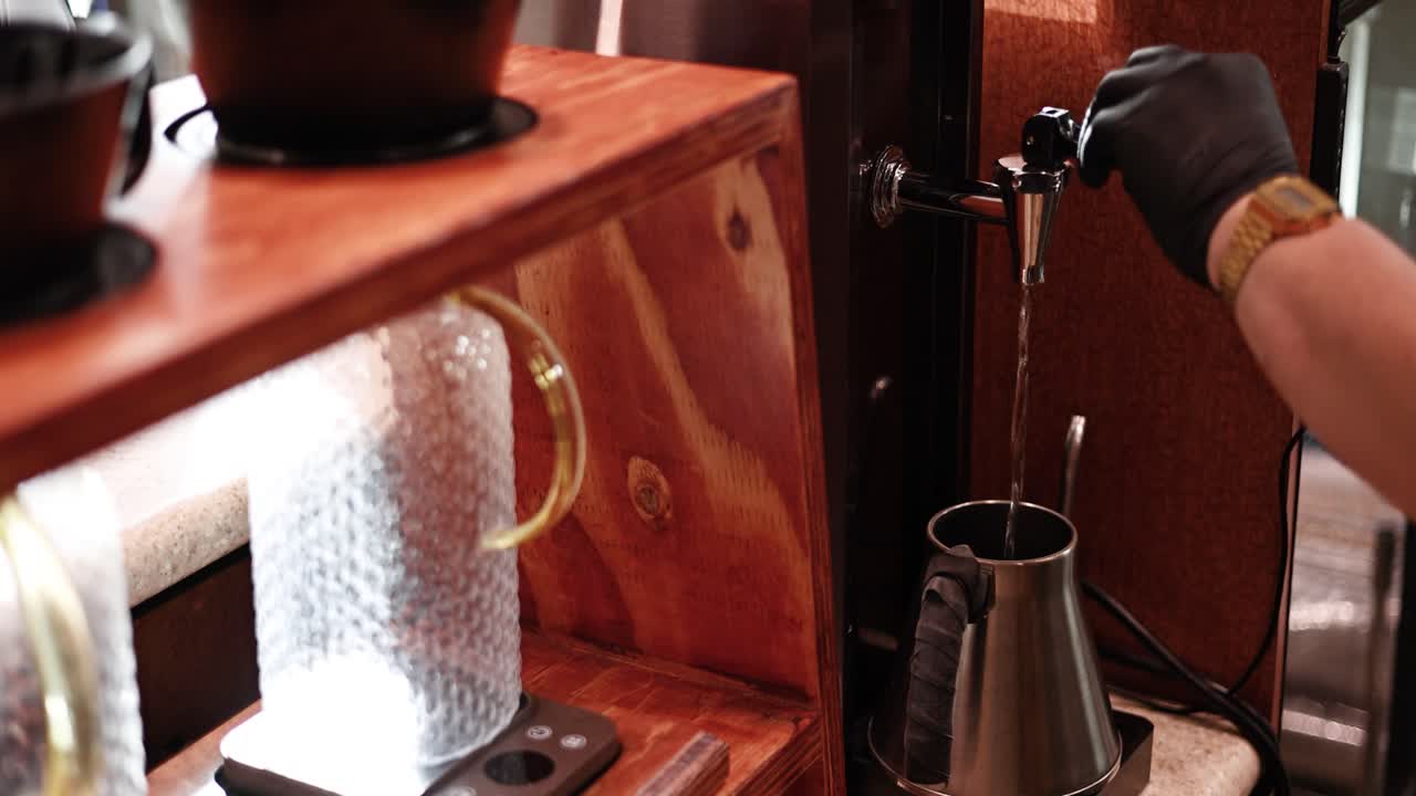 Fremadgående dolly-skud af en barista, der placerer en kedel under en varmtvandshane ved en caféstation – ren kande i rustfrit stål, blød bokeh og flydende arbejdsgang.