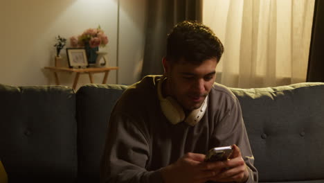 young man wearing wireless headphones spending evening at home sitting on sofa talking on mobile phone 1