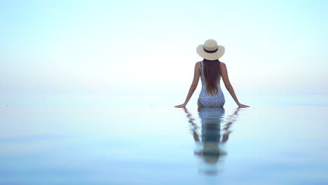 back of sexy woman in swimsuit sitting on infinity pool which merges with tropical sea horizon making heavenly scenery